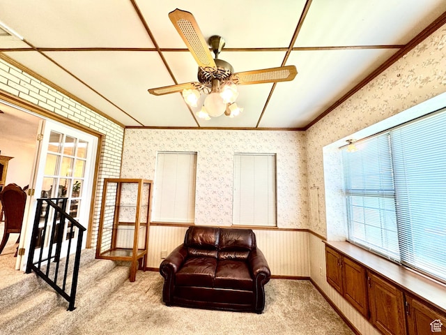 living area featuring healthy amount of natural light, ornamental molding, wallpapered walls, light carpet, and ceiling fan