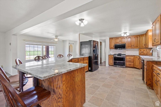 kitchen with appliances with stainless steel finishes, a kitchen breakfast bar, a sink, brown cabinetry, and ceiling fan