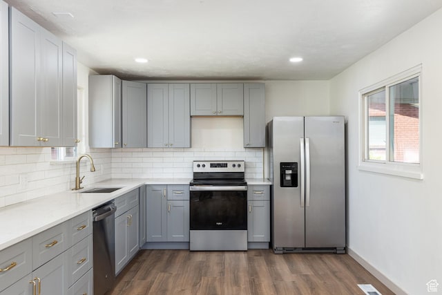 kitchen with appliances with stainless steel finishes, a sink, gray cabinetry, dark wood-style flooring, and tasteful backsplash
