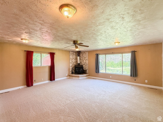 unfurnished living room with a wood stove, a textured ceiling, baseboards, a ceiling fan, and carpet floors