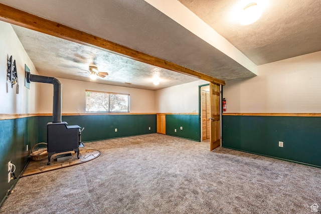 below grade area featuring a wood stove, a textured ceiling, carpet floors, wainscoting, and ceiling fan
