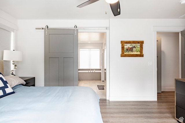 bedroom with light wood-style floors, baseboards, a barn door, ensuite bath, and a ceiling fan