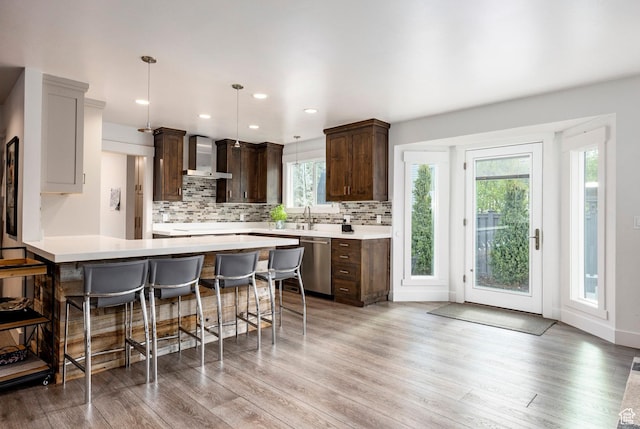 kitchen with dishwasher, wall chimney exhaust hood, dark brown cabinetry, a breakfast bar area, and tasteful backsplash