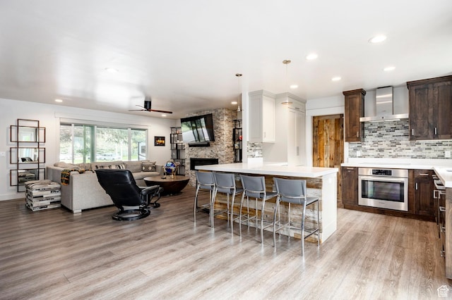 kitchen featuring stainless steel oven, wall chimney exhaust hood, open floor plan, dark brown cabinetry, and light wood finished floors