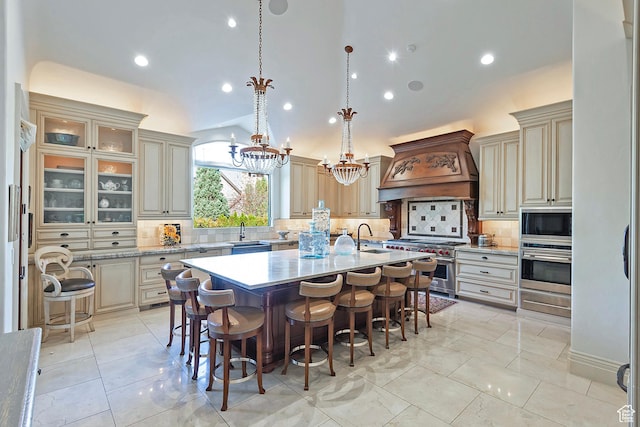 kitchen featuring appliances with stainless steel finishes, premium range hood, cream cabinetry, a kitchen bar, and a kitchen island with sink