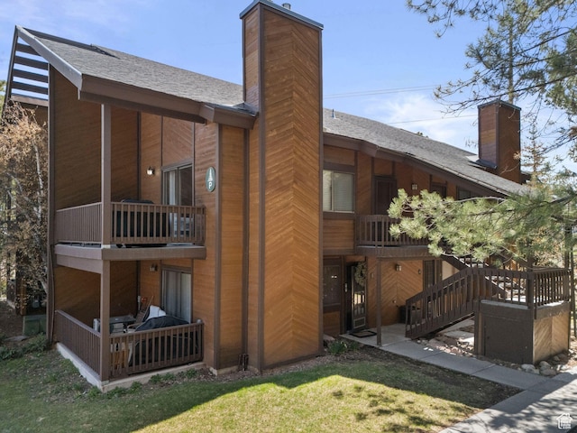 rear view of house featuring a chimney, a balcony, roof with shingles, and a yard