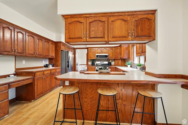 kitchen with appliances with stainless steel finishes, a peninsula, light wood finished floors, brown cabinetry, and a breakfast bar