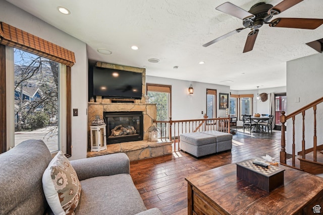 living area featuring wood-type flooring, a ceiling fan, a stone fireplace, a textured ceiling, and stairs