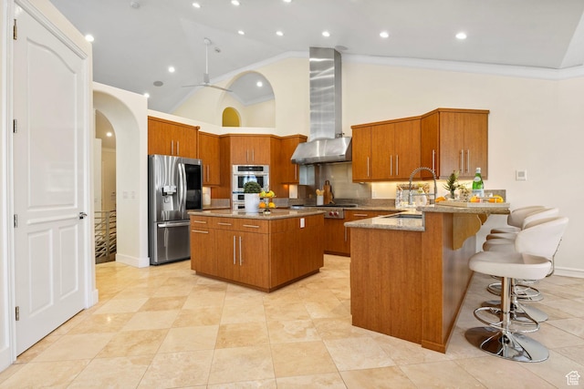 kitchen featuring appliances with stainless steel finishes, wall chimney exhaust hood, a sink, brown cabinets, and high vaulted ceiling