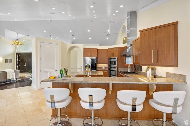 kitchen with appliances with stainless steel finishes, a peninsula, brown cabinetry, light stone counters, and ceiling fan