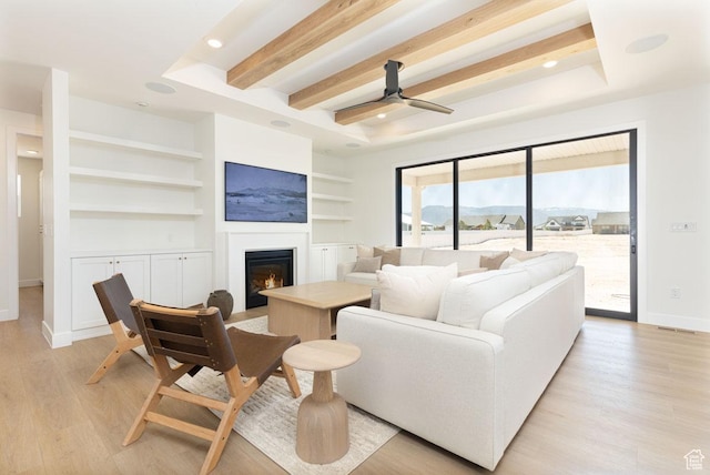 living area with a warm lit fireplace, built in features, baseboards, light wood-style flooring, and beamed ceiling