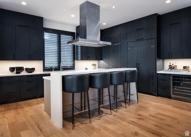 kitchen featuring wine cooler, island exhaust hood, dark cabinetry, a breakfast bar area, and light countertops