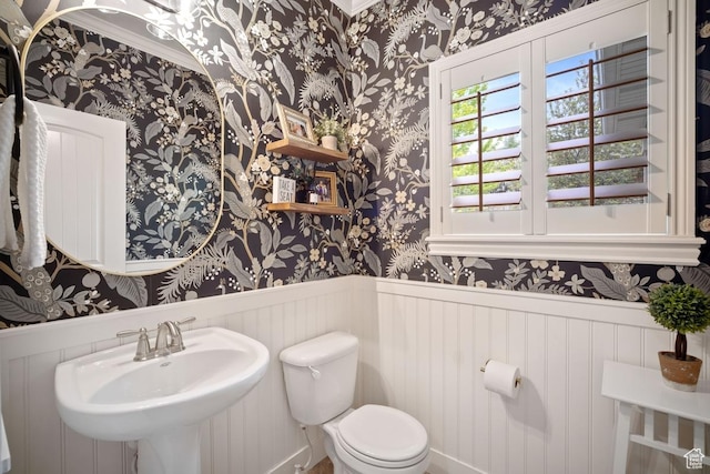 half bathroom with wallpapered walls, a sink, a wainscoted wall, toilet, and crown molding