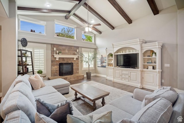 living room with ceiling fan, wood finished floors, beamed ceiling, a stone fireplace, and high vaulted ceiling