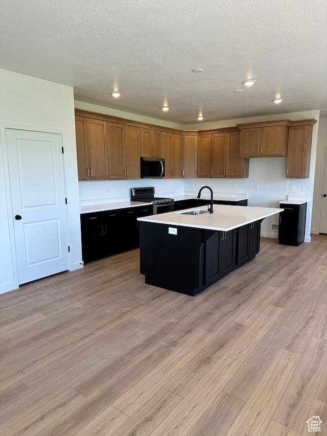 kitchen with stainless steel appliances, a sink, brown cabinets, light wood finished floors, and a kitchen island with sink