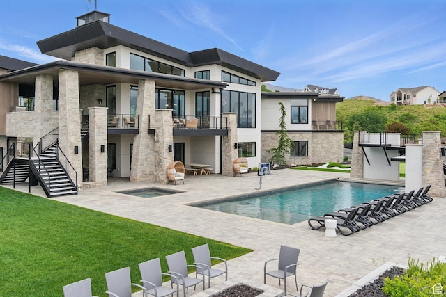 back of house with an in-ground hot tub, a balcony, a patio, stone siding, and a lawn