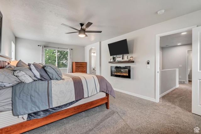 carpeted bedroom with a glass covered fireplace, arched walkways, baseboards, a ceiling fan, and ensuite bath