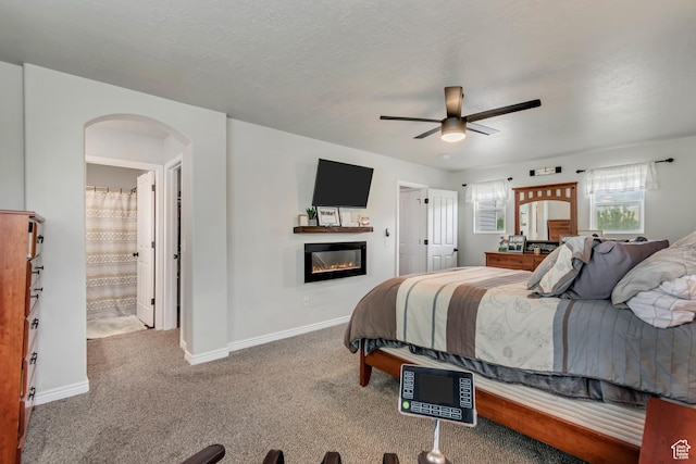 bedroom featuring carpet floors, a glass covered fireplace, arched walkways, baseboards, and ceiling fan