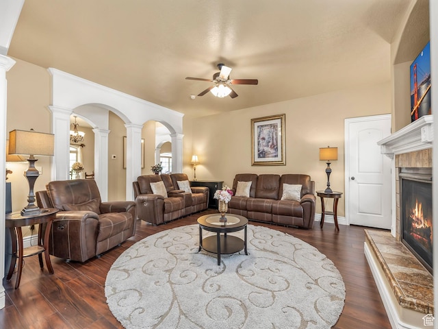 living area featuring decorative columns, a ceiling fan, a tiled fireplace, arched walkways, and dark wood-style floors