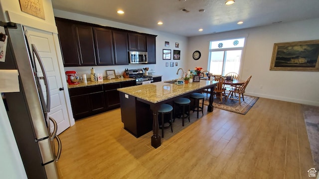 kitchen featuring stainless steel appliances, a breakfast bar, light wood finished floors, an island with sink, and light stone counters