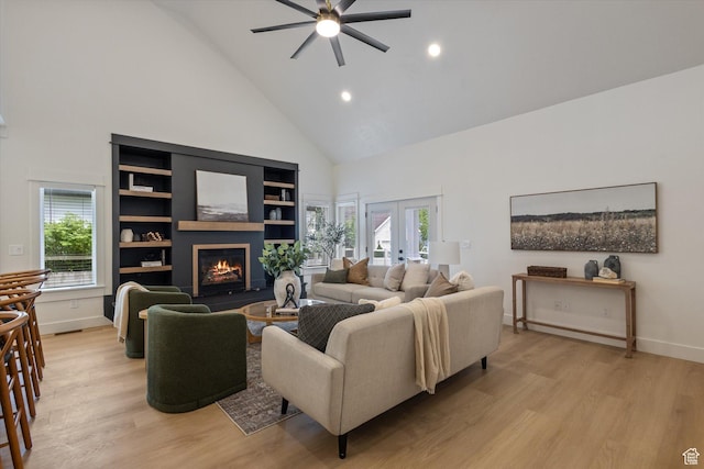 living area featuring high vaulted ceiling, ceiling fan, light wood-type flooring, a glass covered fireplace, and healthy amount of natural light