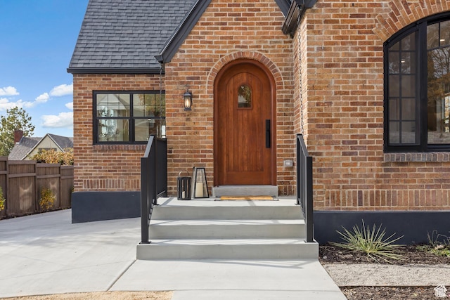entrance to property with brick siding, fence, and roof with shingles