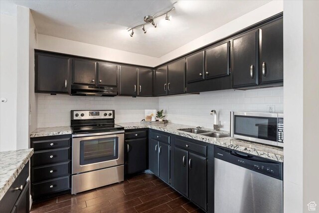 kitchen with appliances with stainless steel finishes, dark wood finished floors, a sink, under cabinet range hood, and backsplash