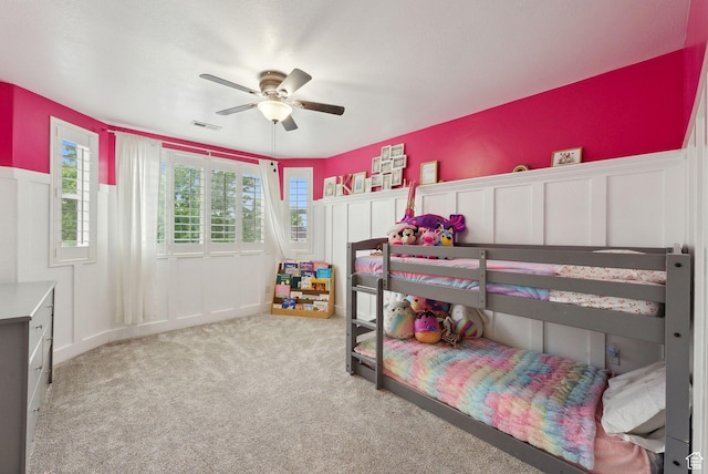 carpeted bedroom with a decorative wall, wainscoting, and a ceiling fan