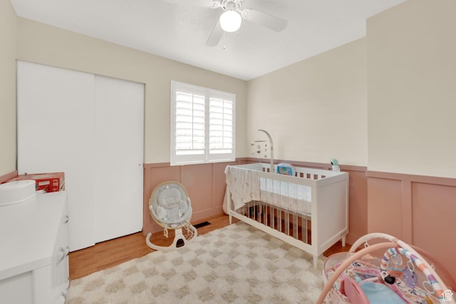 bedroom with wood finished floors, a wainscoted wall, a closet, a ceiling fan, and a nursery area