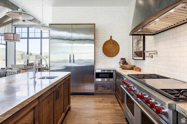 kitchen with high end appliances, a sink, wall chimney range hood, light wood finished floors, and backsplash