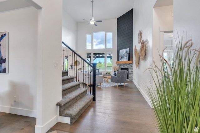 stairs with ceiling fan, baseboards, hardwood / wood-style floors, a stone fireplace, and a towering ceiling
