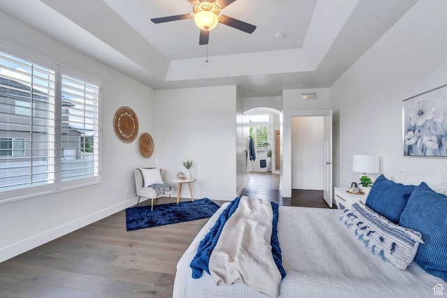 bedroom featuring arched walkways, a raised ceiling, wood finished floors, baseboards, and ceiling fan