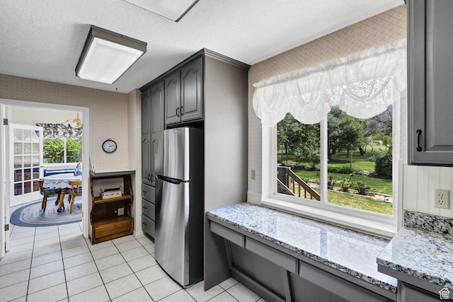 kitchen featuring freestanding refrigerator, gray cabinetry, light stone countertops, light tile patterned floors, and wallpapered walls
