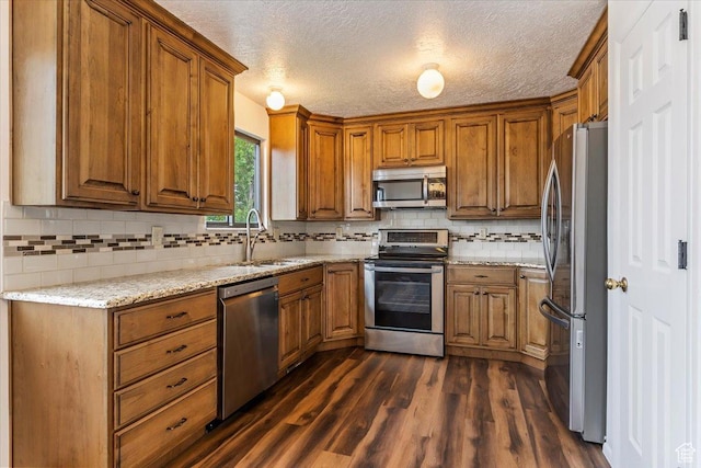 kitchen with appliances with stainless steel finishes, a sink, brown cabinetry, dark wood finished floors, and tasteful backsplash