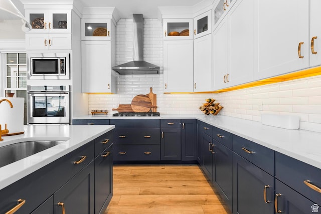 kitchen with stainless steel appliances, wall chimney exhaust hood, light wood-style floors, white cabinets, and tasteful backsplash