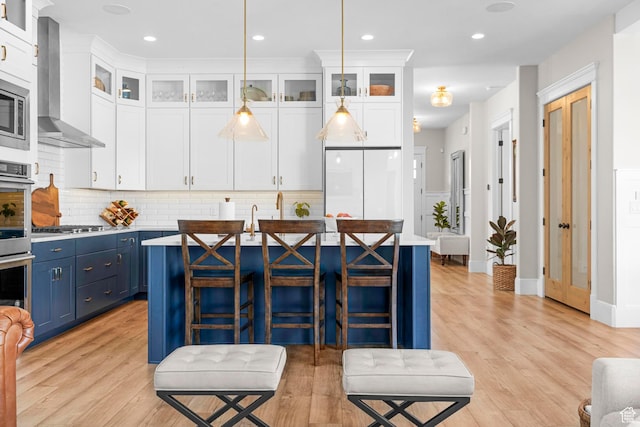 kitchen featuring blue cabinetry, wall chimney exhaust hood, a kitchen island with sink, white cabinetry, and decorative backsplash