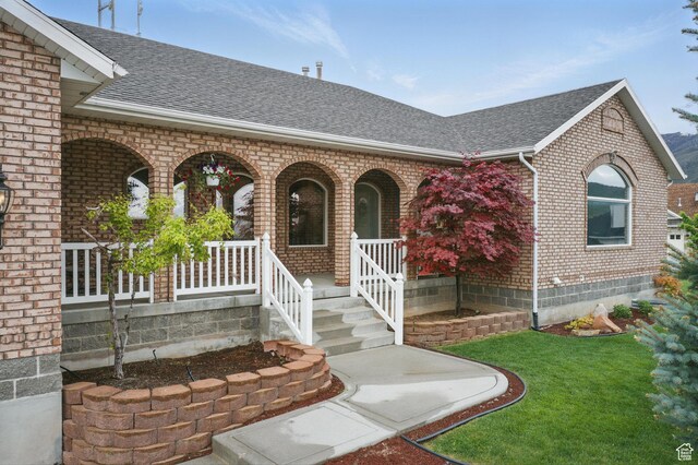 doorway to property with brick siding, roof with shingles, a porch, and a yard