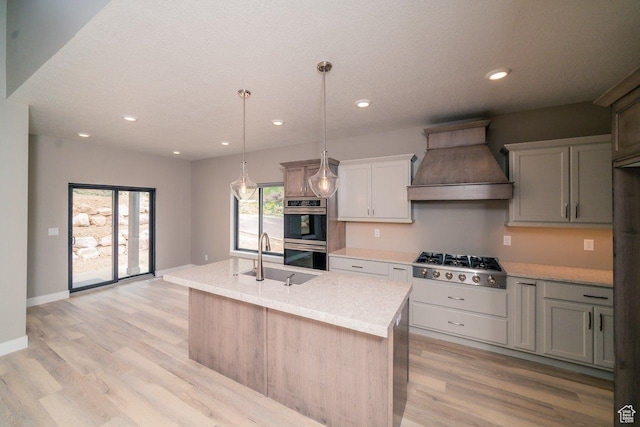 kitchen with custom range hood, stainless steel appliances, a sink, light wood-style floors, and a kitchen island with sink
