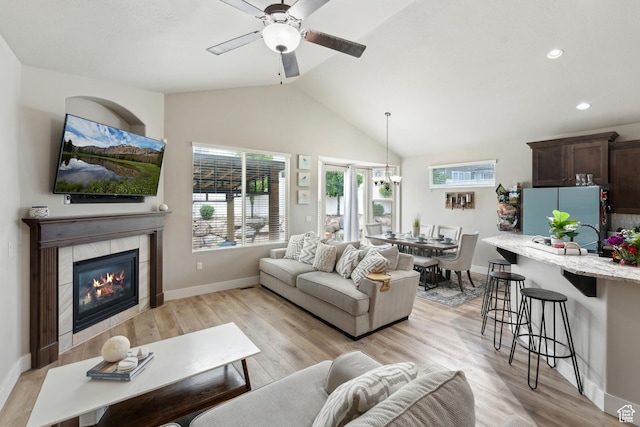 living room with a ceiling fan, light wood-style floors, vaulted ceiling, a tiled fireplace, and baseboards