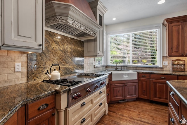 kitchen with a sink, custom exhaust hood, stainless steel gas cooktop, backsplash, and dark stone counters