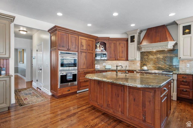 kitchen with custom range hood, stainless steel appliances, glass insert cabinets, a center island with sink, and recessed lighting