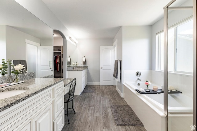 bathroom featuring wood finished floors, two vanities, a bath, baseboards, and a spacious closet