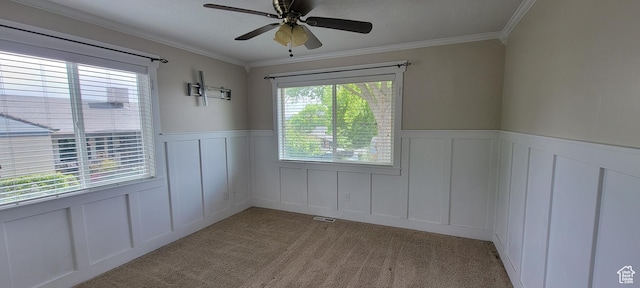 empty room featuring a decorative wall, wainscoting, a ceiling fan, light colored carpet, and crown molding