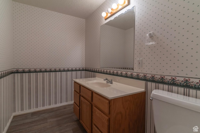 bathroom with wallpapered walls, wood finished floors, a wainscoted wall, toilet, and vanity