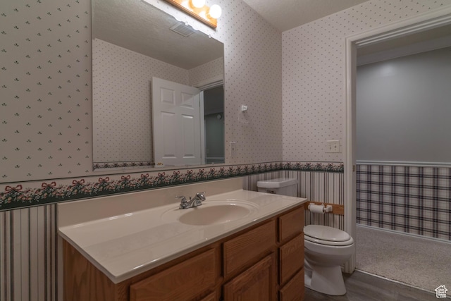 bathroom featuring wallpapered walls, toilet, vanity, wood finished floors, and wainscoting