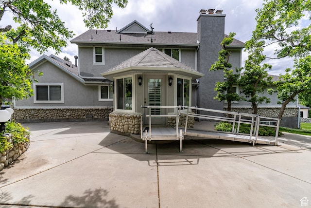 rear view of house with stucco siding, a chimney, a patio, stone siding, and roof with shingles