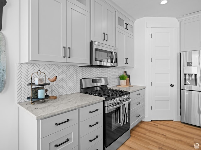 kitchen featuring appliances with stainless steel finishes, light wood-style flooring, decorative backsplash, light stone counters, and glass insert cabinets