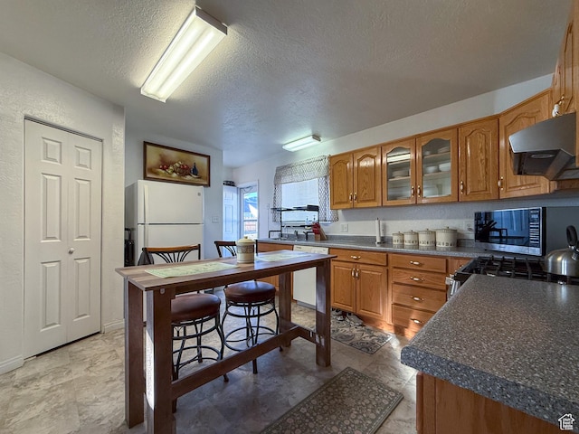 kitchen featuring range hood, a textured ceiling, brown cabinetry, appliances with stainless steel finishes, and dark countertops