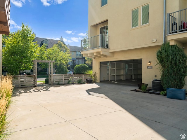 view of patio featuring a balcony, concrete driveway, a garage, fence, and central air condition unit