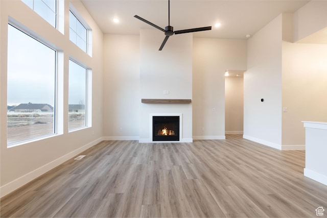 unfurnished living room featuring a warm lit fireplace, light wood-style flooring, baseboards, a ceiling fan, and a high ceiling